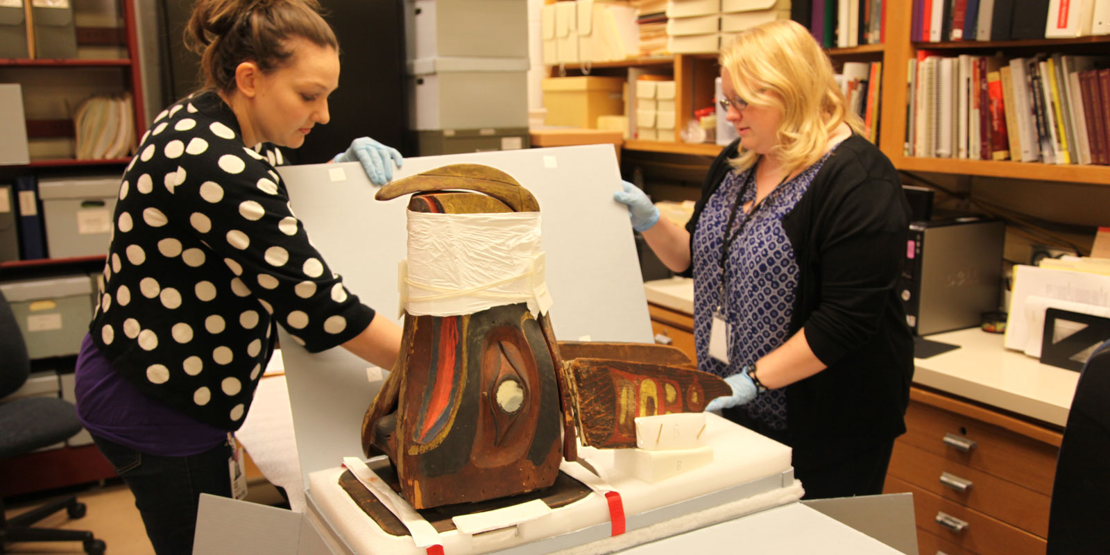 Two woman carefully unpack the native art mask from a box