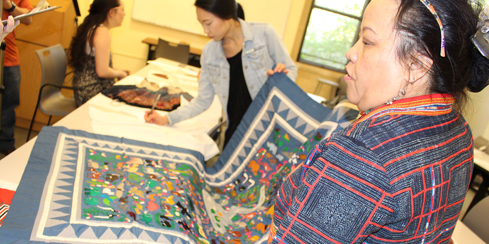 two women hold a hmong blanket while one takes notes on its details