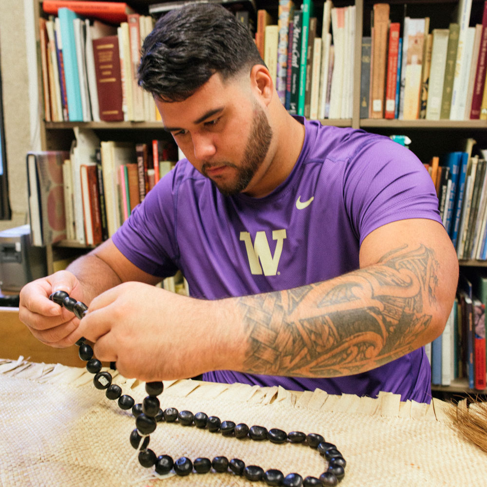 A male student in purple UW Husky tee shirt holding a beaded necklace alongside a woven Samoan rug 