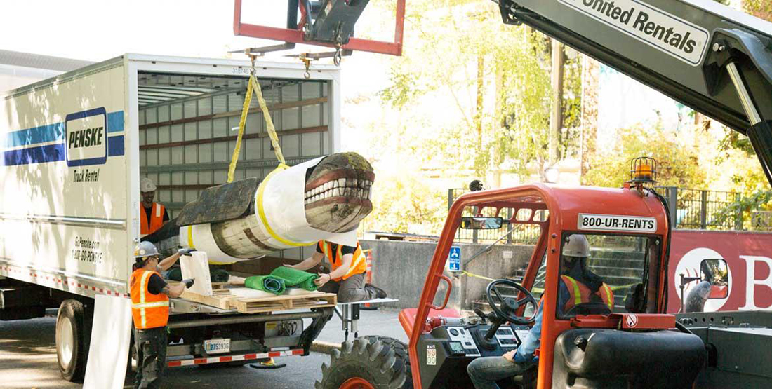A whale carving is carefully placed inside a truck by a crane