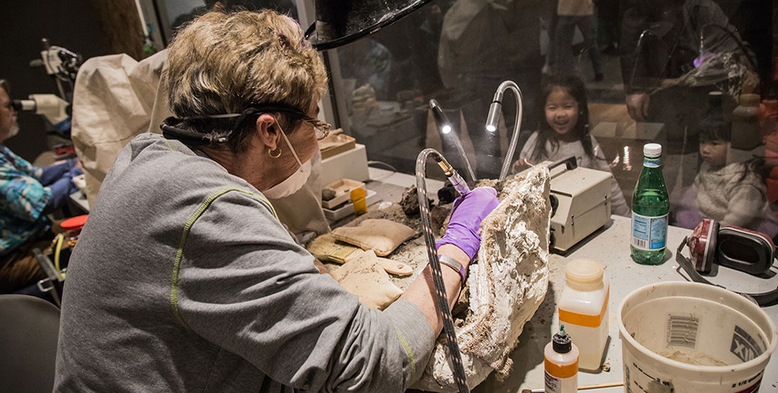 A woman preparing a fossil smiles at two young girls behind the glass