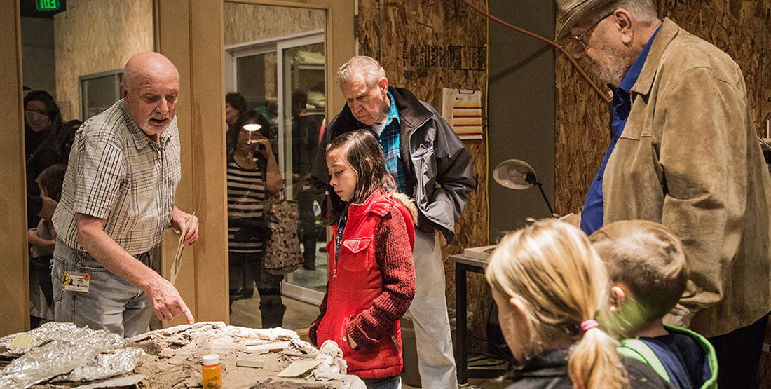 A man shows a multi-generational group of visitors the fossil he's preparing