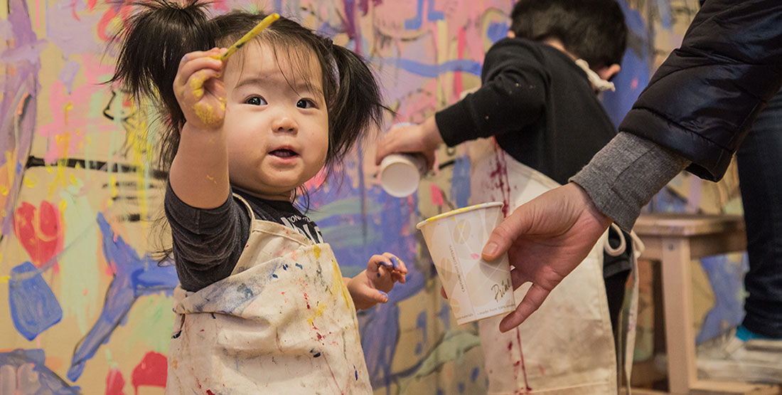 A toddler girl holds up a paintbrush covered in yellow paint