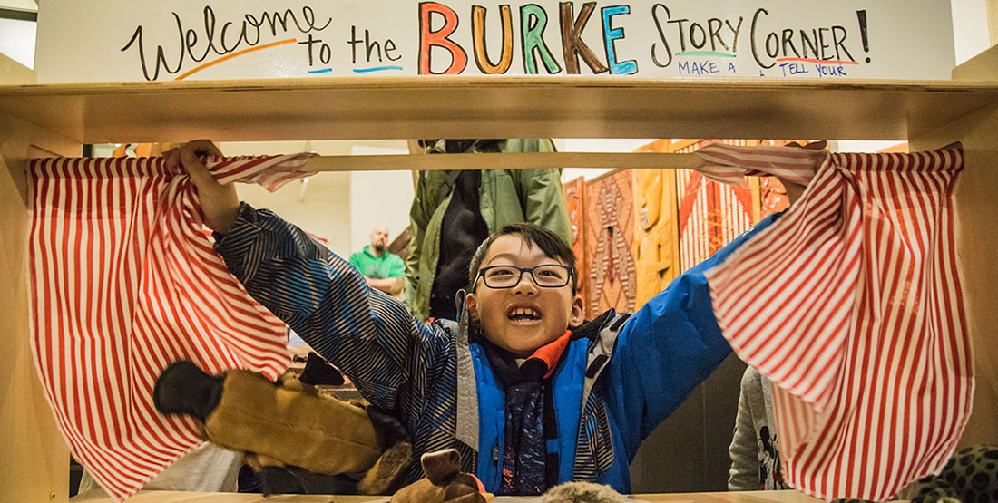 A young boy opens the curtain of a puppet story corner space with a big smile