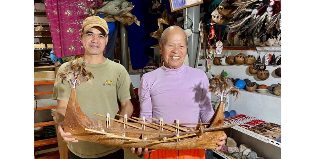 Two men stand side by side holding a model canoe in the style of Tatala that they made together.