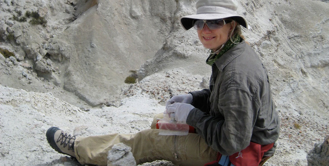 A female researcher wearing a hat and sunglasses sits on the rocky ground