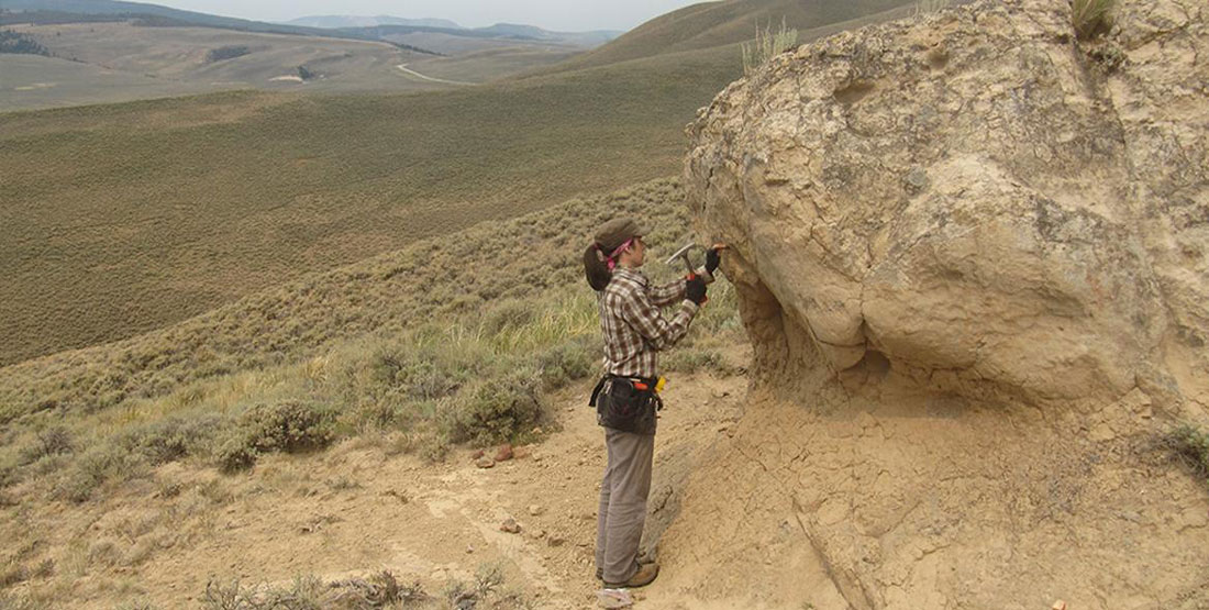 a woman reaches up to reove a sample from a rock outcrop