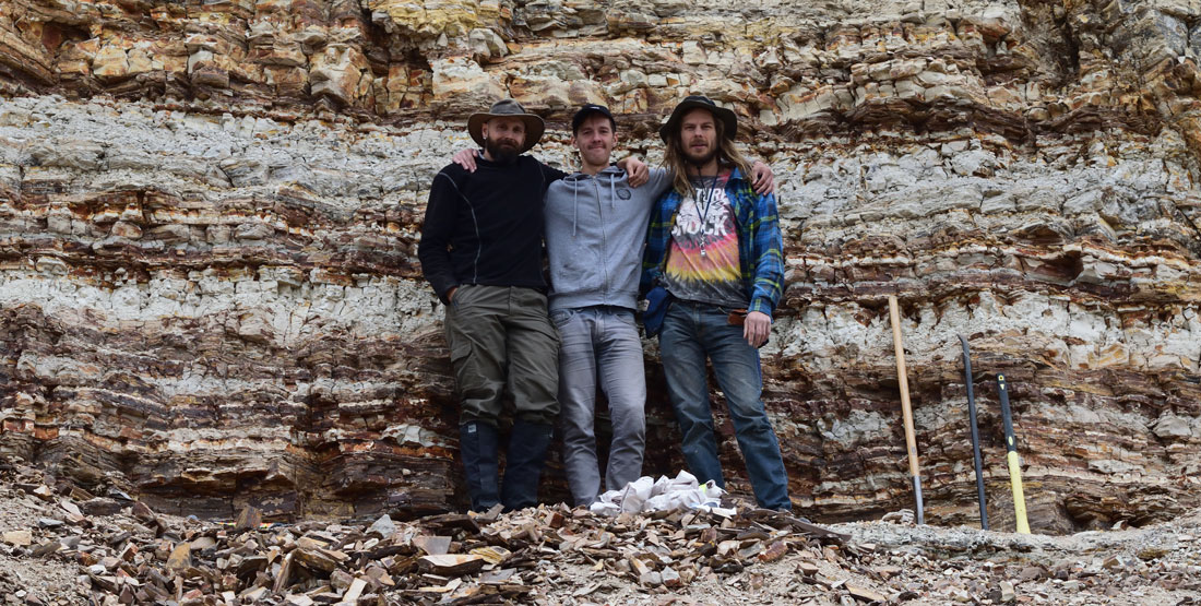three young men stand in front of a rock wall showing many stratigraphic layers