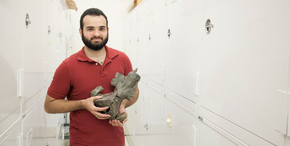 a young man stands within a row in the paleontology collections room and holds a fossil whale skull