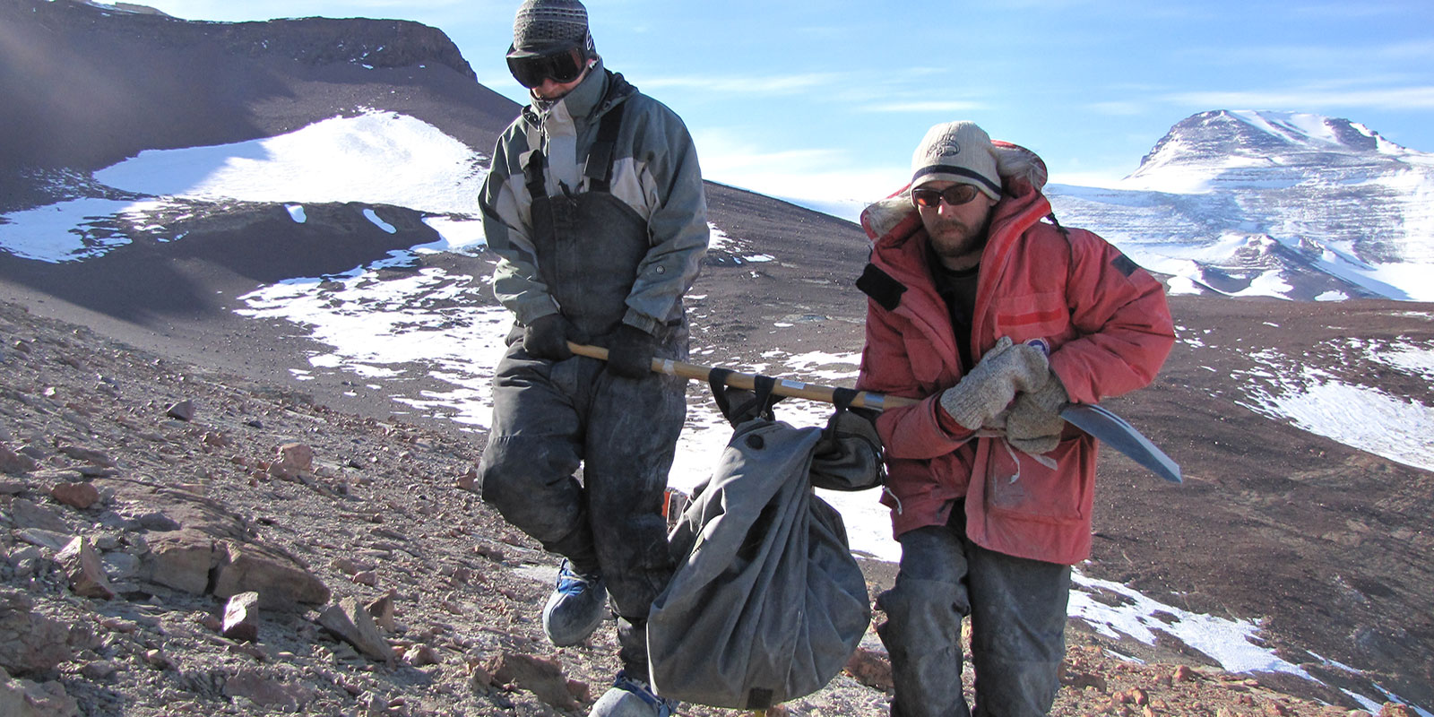 Two men use a shovel to carry a heavy fossil encased in rock out of the Antarctic landscape