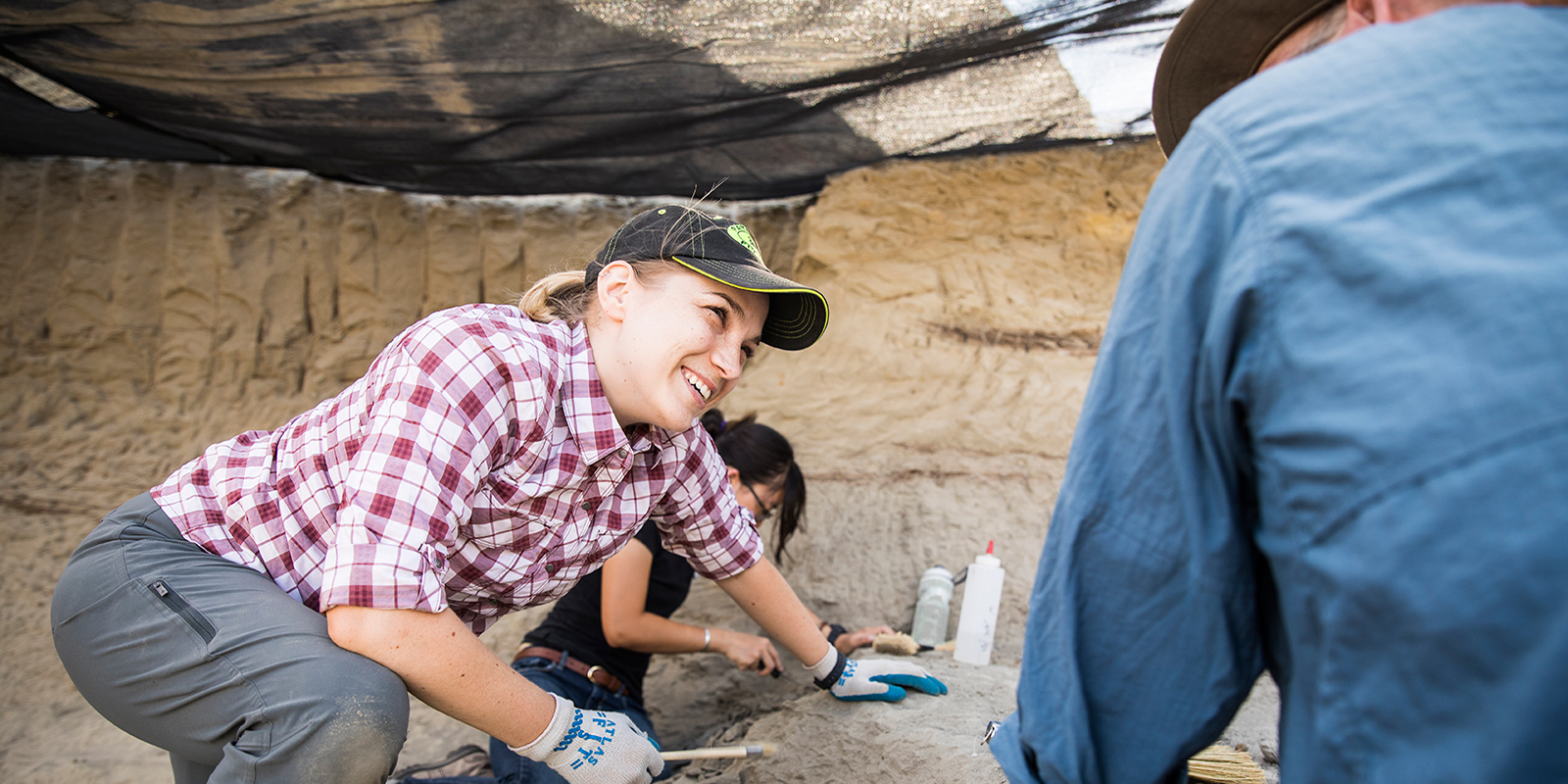 A woman in dirty working clothes smiling at another person under a canopy on a paleontology dig