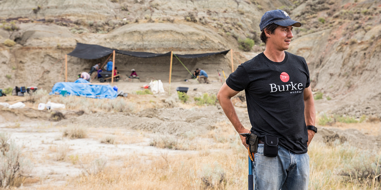 A palentologist with a Burke Museum tee shirt standing in front of a paleontology dig site
