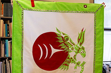 A button-blanket with green, white and red colors includes the Burke Museum logo and an eagle breaking out of chains