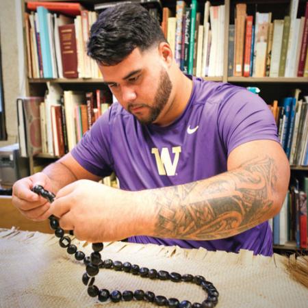 A male student in purple UW Husky tee shirt holding a beaded necklace alongside a woven Samoan rug 