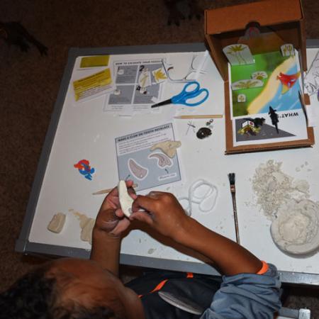a child makes a clay tooth necklace in the fossil finder fun pack box from the Burke Museum