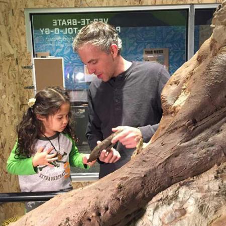 a man shows a girl a fossil while the stand in front of the t.rex skull
