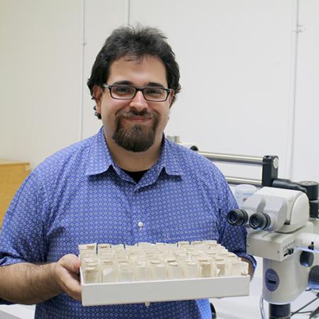 A male researcher holds a tray of tubes and stands next to a microscope