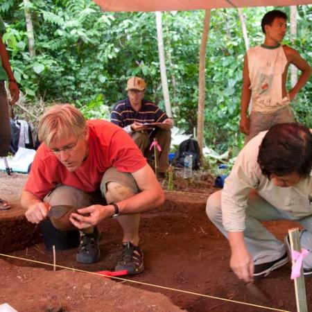 Two men kneel in an archaeological dig site and closely examine sediment for artifacts