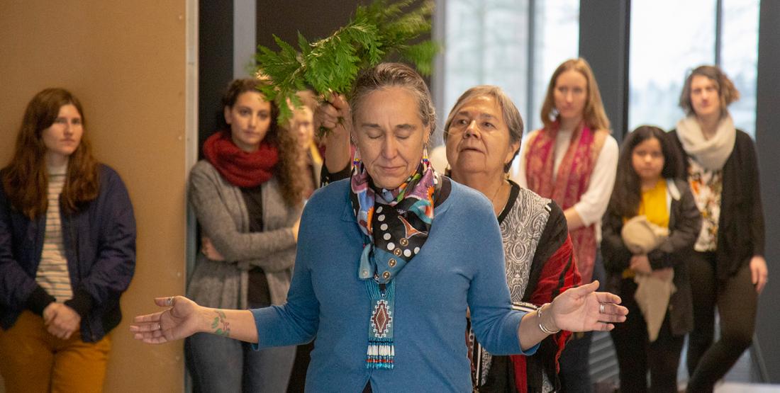 a woman stands with open arms as cedar is brushed over her head