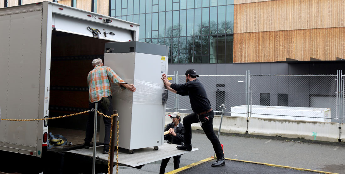three men roll a giant freezer onto the back of a box truck
