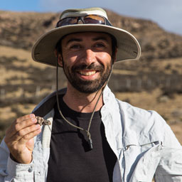 a young man wears a hat and holds a lizard in the desert