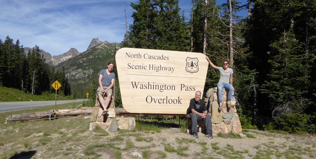 Three people pose for a photo in front of a sign that says North Cascades Scenic Highway, Washington Pass Outlook next to the road