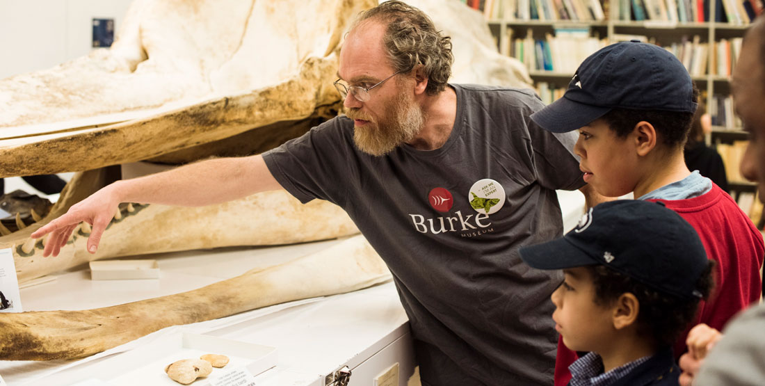 a man points to a feature on a whale skull as 3 children listen intently