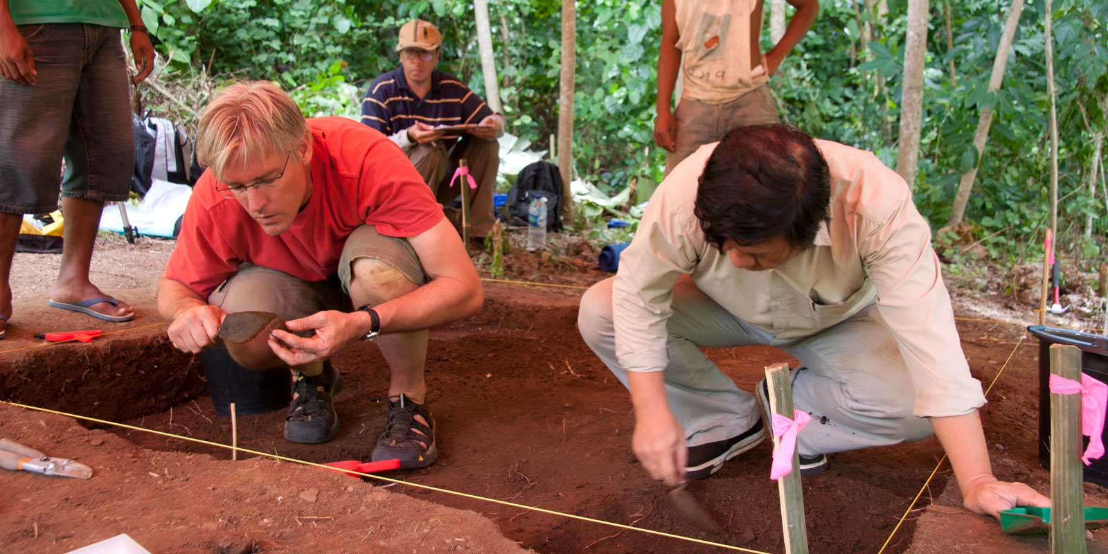 Two men kneel in an archaeological dig site and closely examine sediment for artifacts