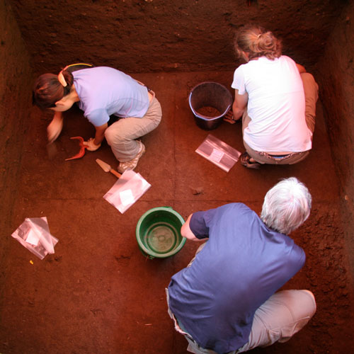 looking down into a pit where two women and a man kneel down digging 