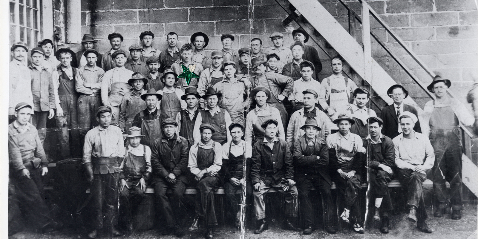A 1920 photo of a large group of men in workwear posing for a photo in front of stairs and a brick wall
