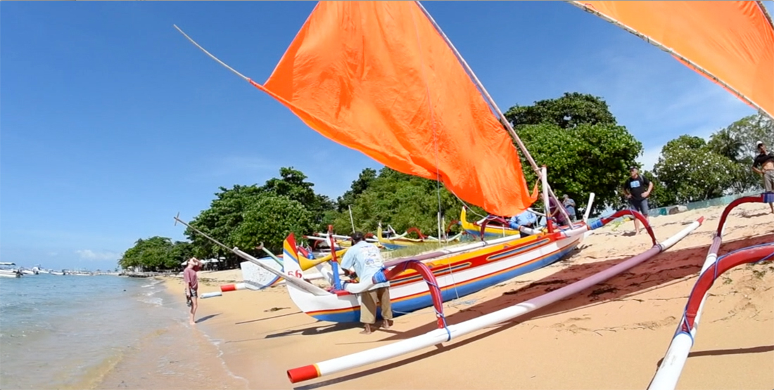 A boat with outriggers and a large open sail is pushed into the water 