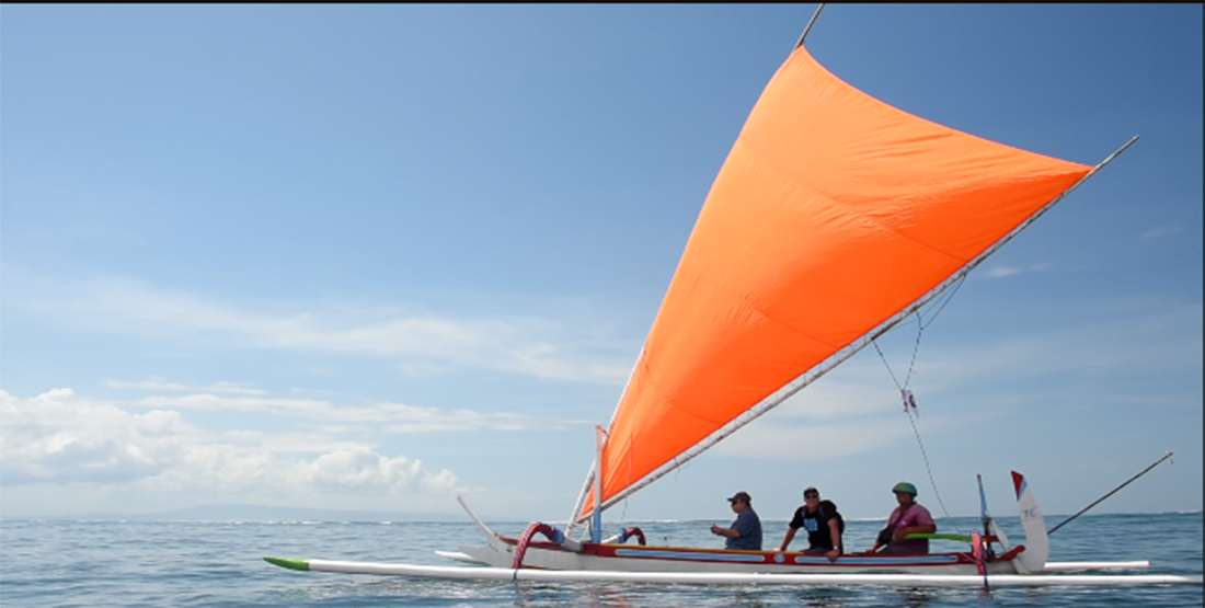 Three people sail a boat with outriggers on the water