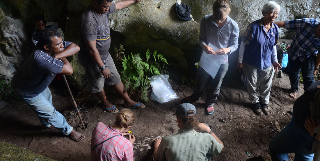 a group of people stand and watch as two researchers kneel on the ground and work