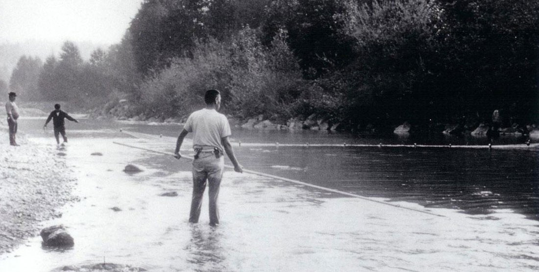 black and white photo of a man standing with a large fishing spear out over the water