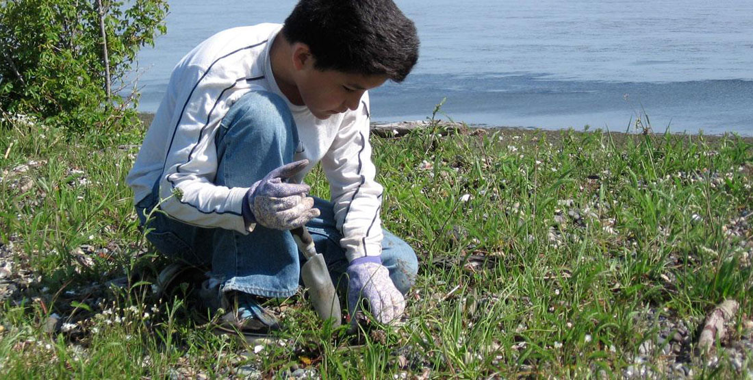 a boy uses a shovel to dig wild onions near the water