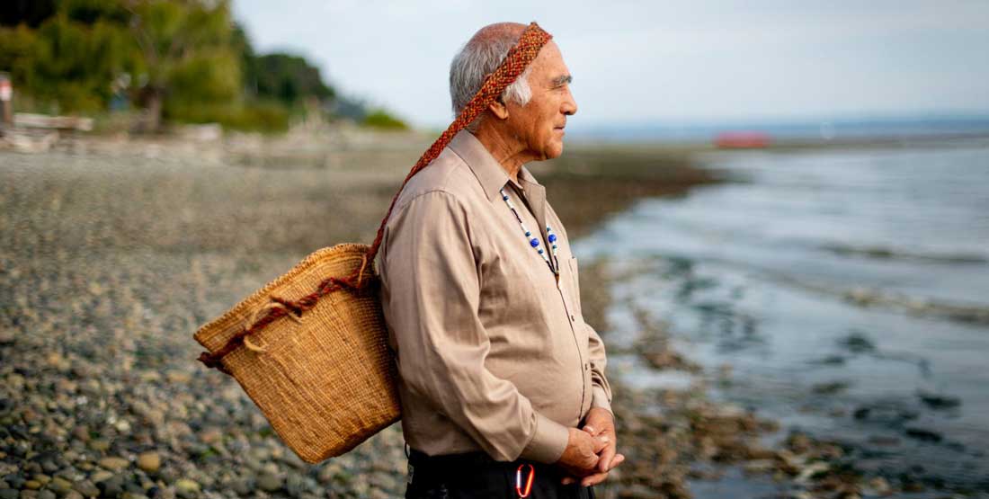 ed carriere stands on the shore of puget sound looking out at the water while holding a basket on his back