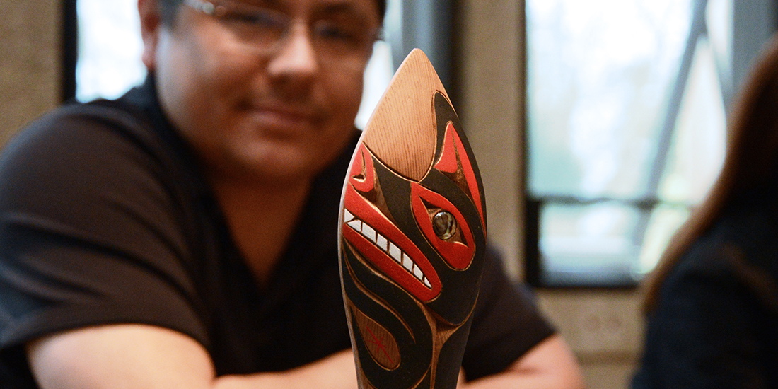 a man sits behind a carved and painted paddle rattle