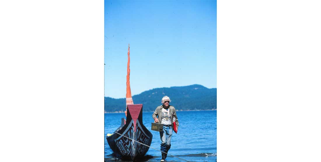 bill holm stands on the shore with a canoe to his left and a bright blue sky in the background