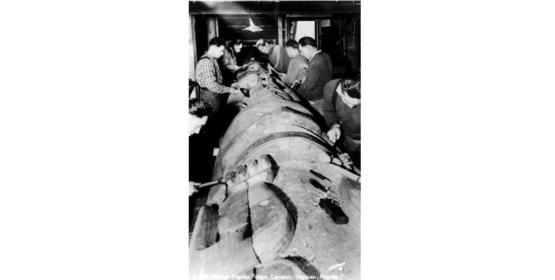 a black and white photo of people carving a wooden totem in alaska