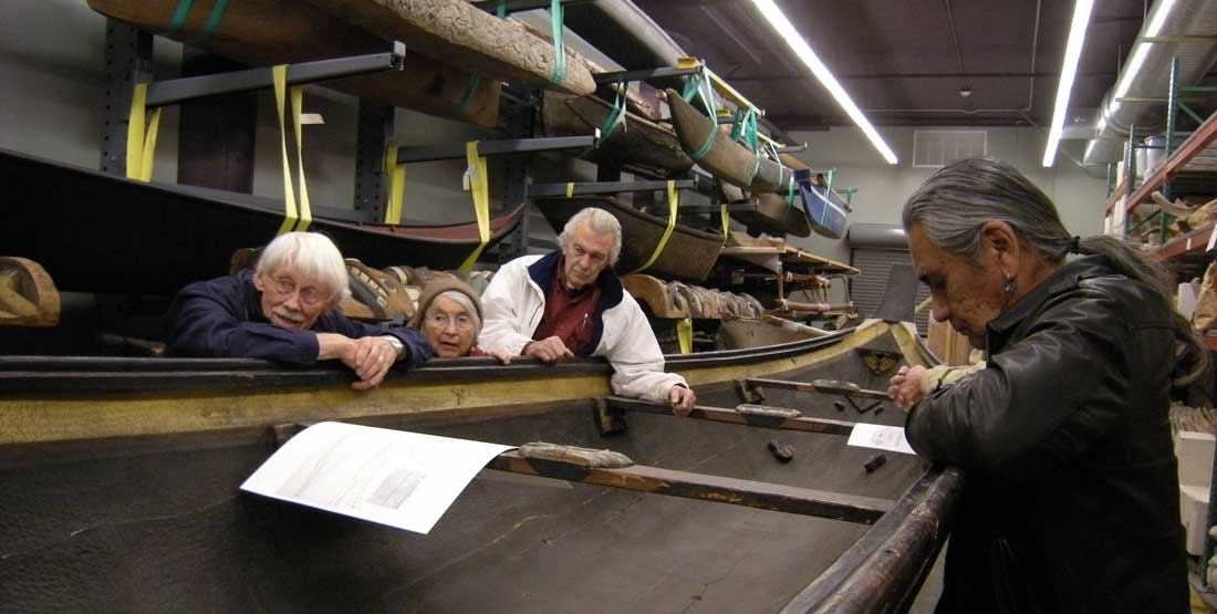 people stand around a carved wood canoe inside a storage building