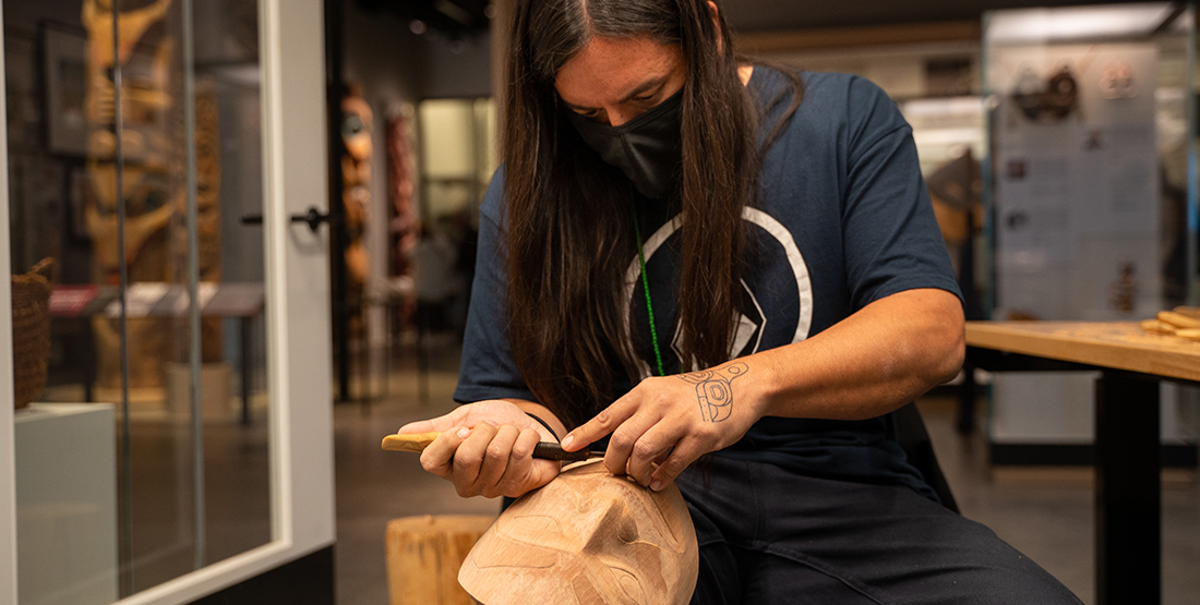 a man carves a mask while sitting in a native art gallery
