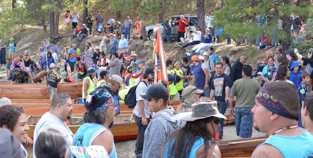 A large crowd of people standing near canoes on the shore of a river