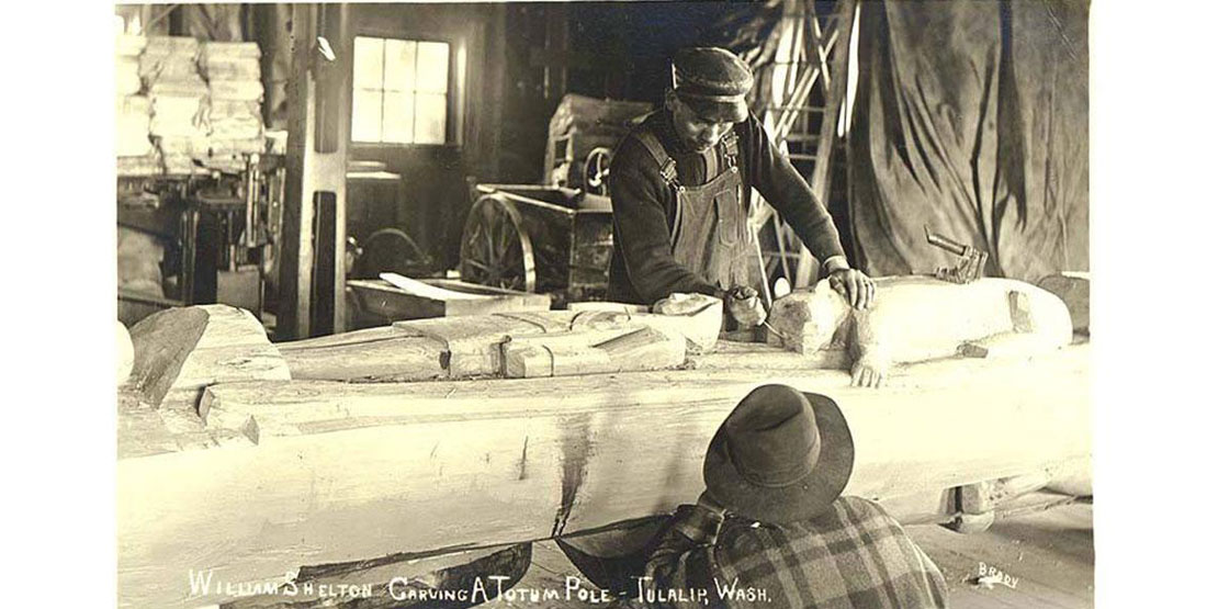 A old photograph of a man carving a totem pole in a workshop while another man looks on