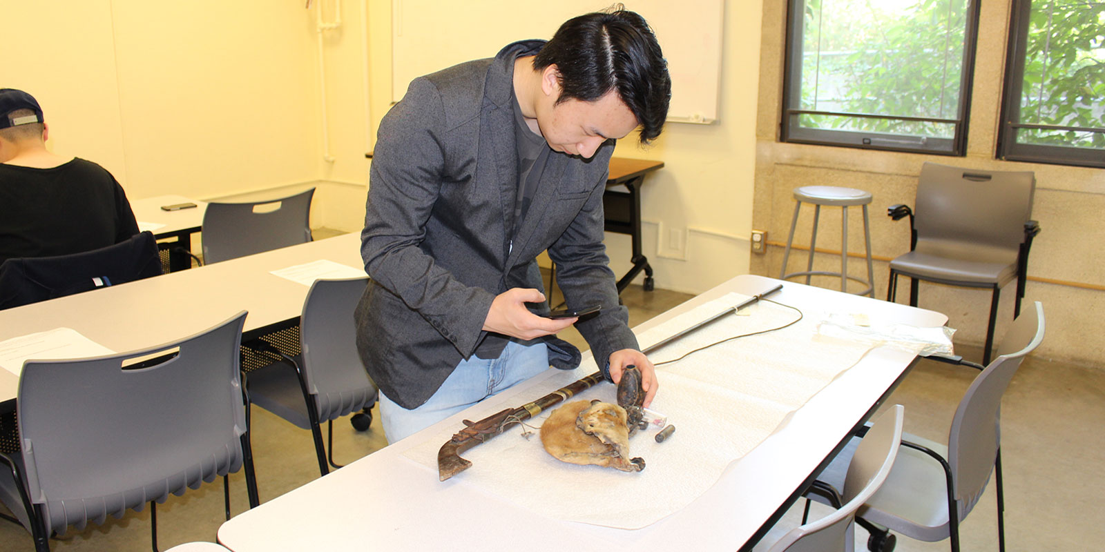 a man holds a cell phone while taking photos of Hmong objects sitting on a table