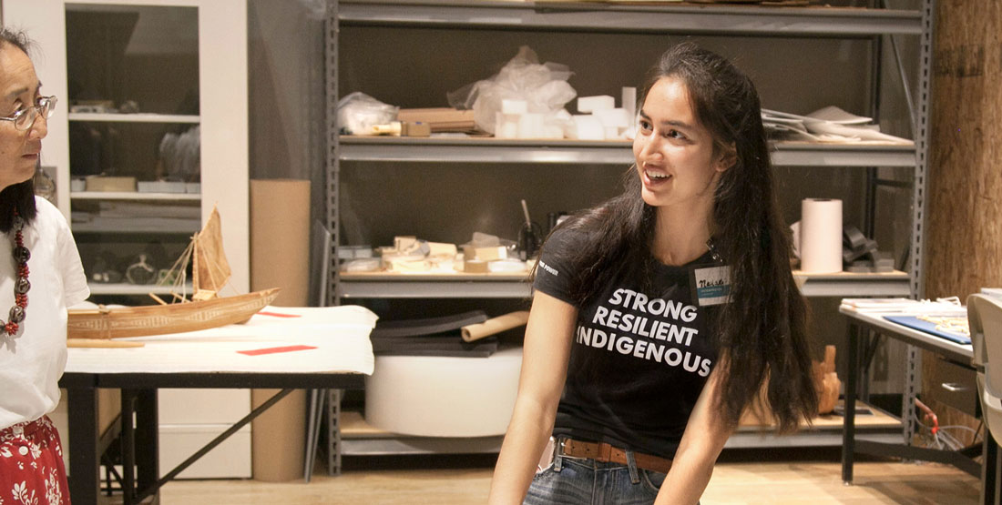 a young woman presents an object in a workroom while wearing a shirt that reads "Strong, Resilient, Indigenous."