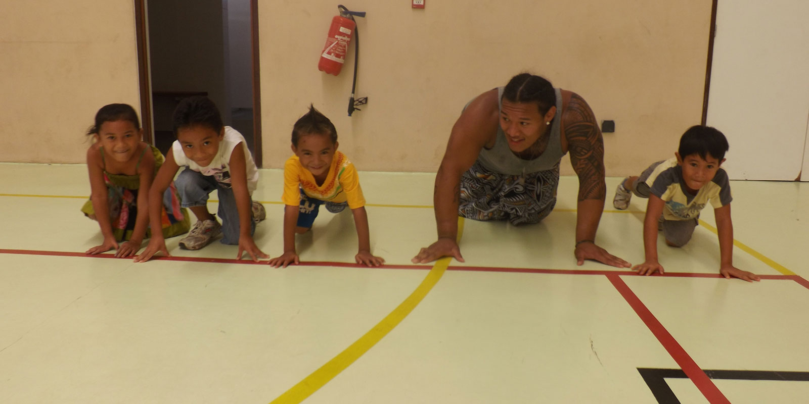 a young man crouches on the ground of a gymnasium with several children