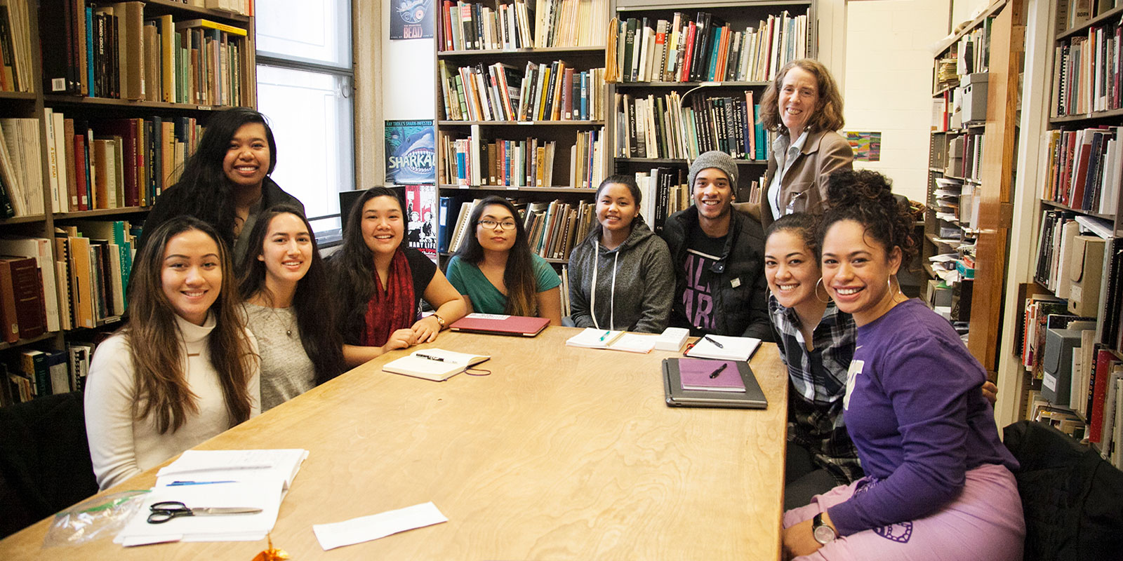 a large group of young women sit around a table in the Burke collections area with their professor 