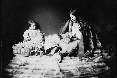 black and white photograph of a woman weaving a basket facing up with a child watching