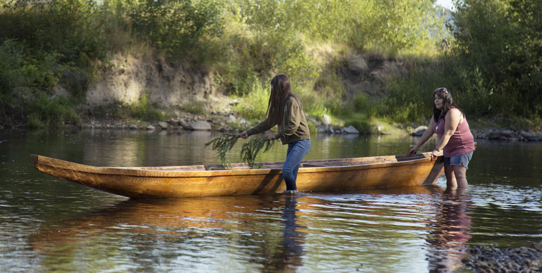 two women place cedar branches in a carved canoe floating on the water