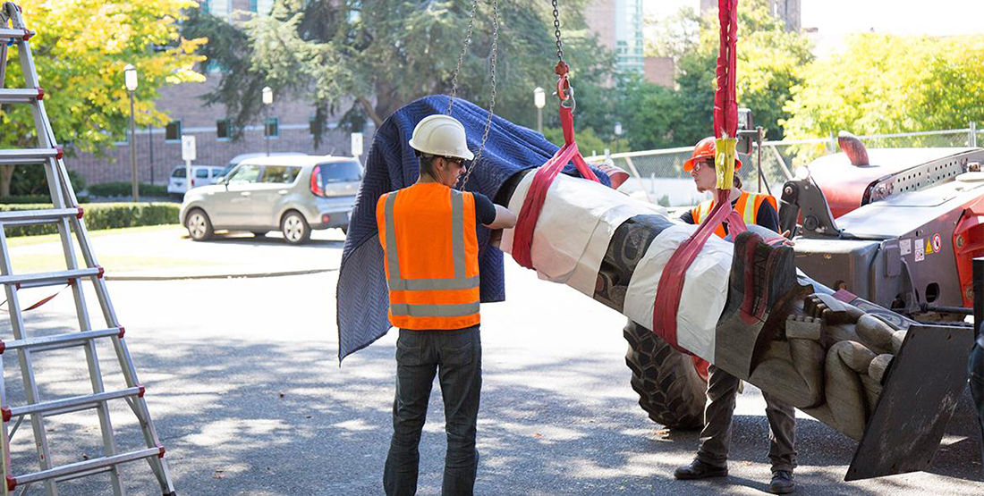 A man places a blanket over a totem pole as it is hoisted by a crane